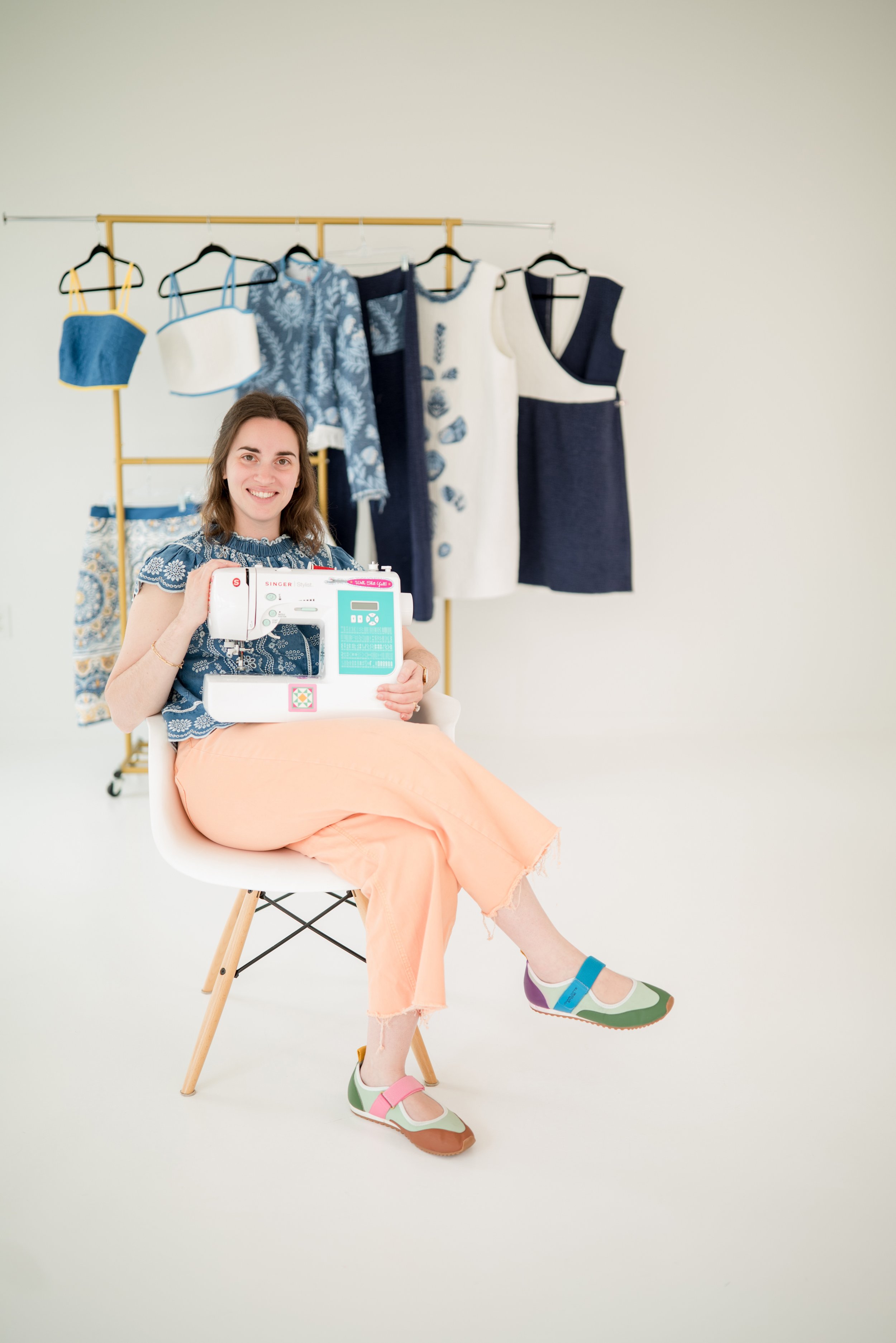 Brigitte of Speedy B Stitchery seated at a sewing machine in front of a rack of handmade garments